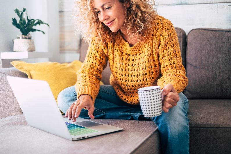 a woman in a yellow sweater holds a coffee mug and smiles as she uses a laptop on the coffee table