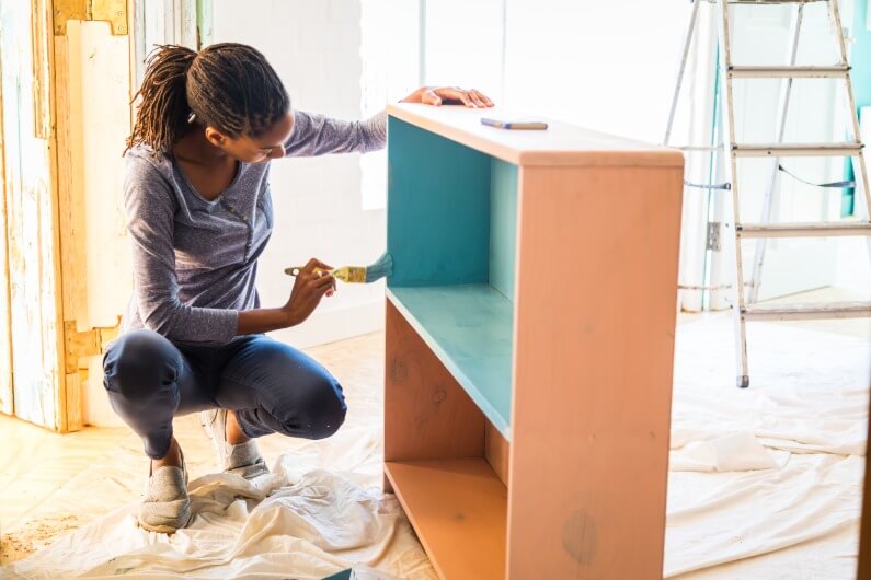 A young homeowner kneels down to paint a refurbished piece of furniture for her new home.
