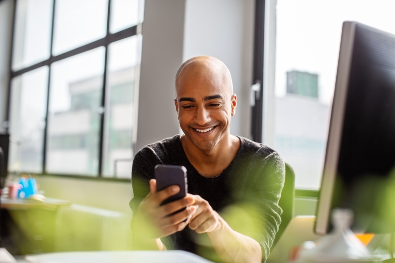 a man smiles at a phone in his hand while sitting at a desk with a monitor