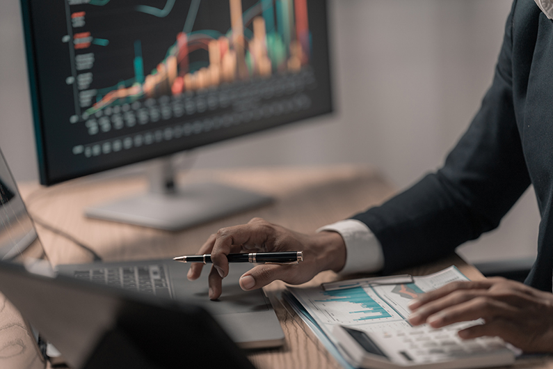 Person working at a desk with a laptop and financial documents, holding a pen while analyzing charts and graphs. A computer monitor in the background displays colorful bar and line graphs on a dark screen.