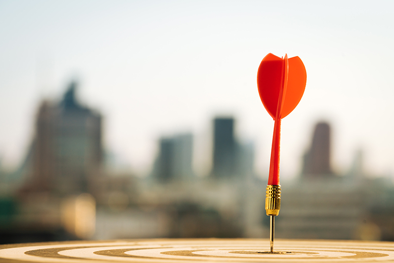Close-up of a red dart standing upright in the bullseye of a dartboard, with a blurred city skyline in the background.