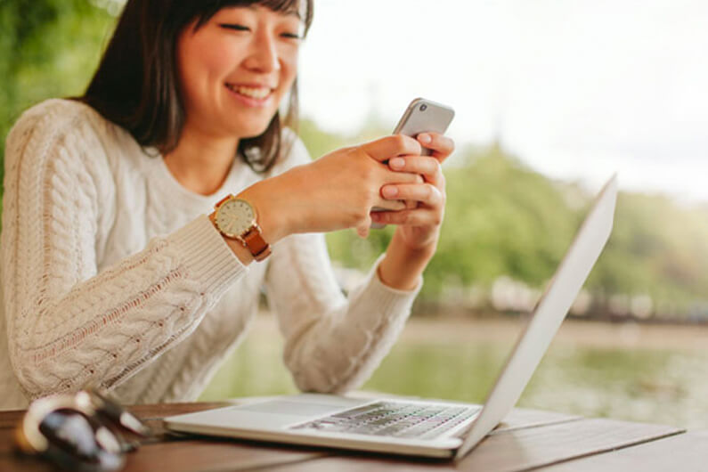 a woman smiles at a phone in her hands while sitting in front of a laptop on a table with a lake in the background